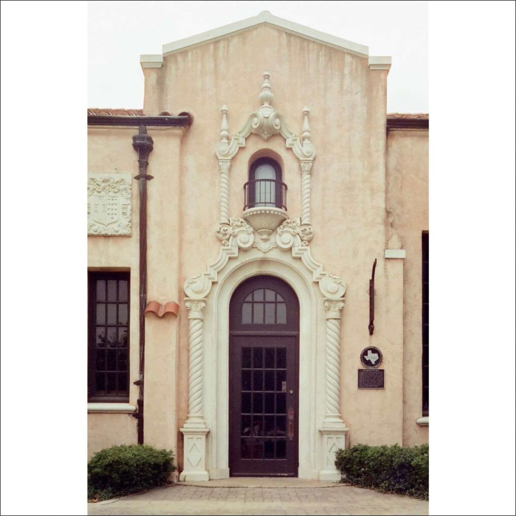 The image shows a beige stucco building with an ornate white stone archway framing a dark brown arched door.