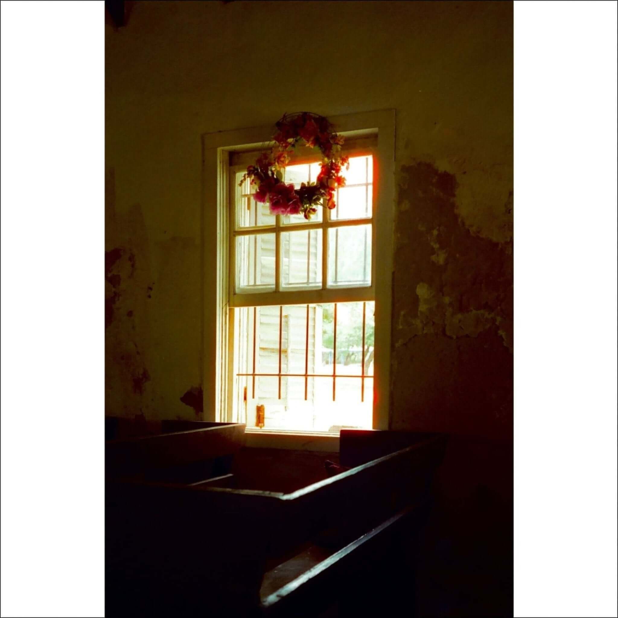 A floral wreath with red and pink blossoms hangs above a window in an old, sunlit room.