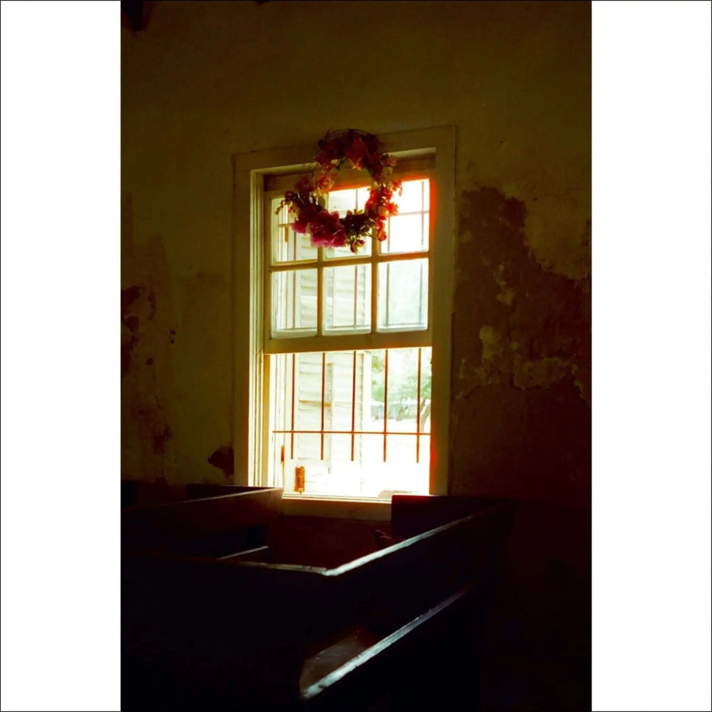 A floral wreath with red and pink blossoms hangs above a window in an old, sunlit room.