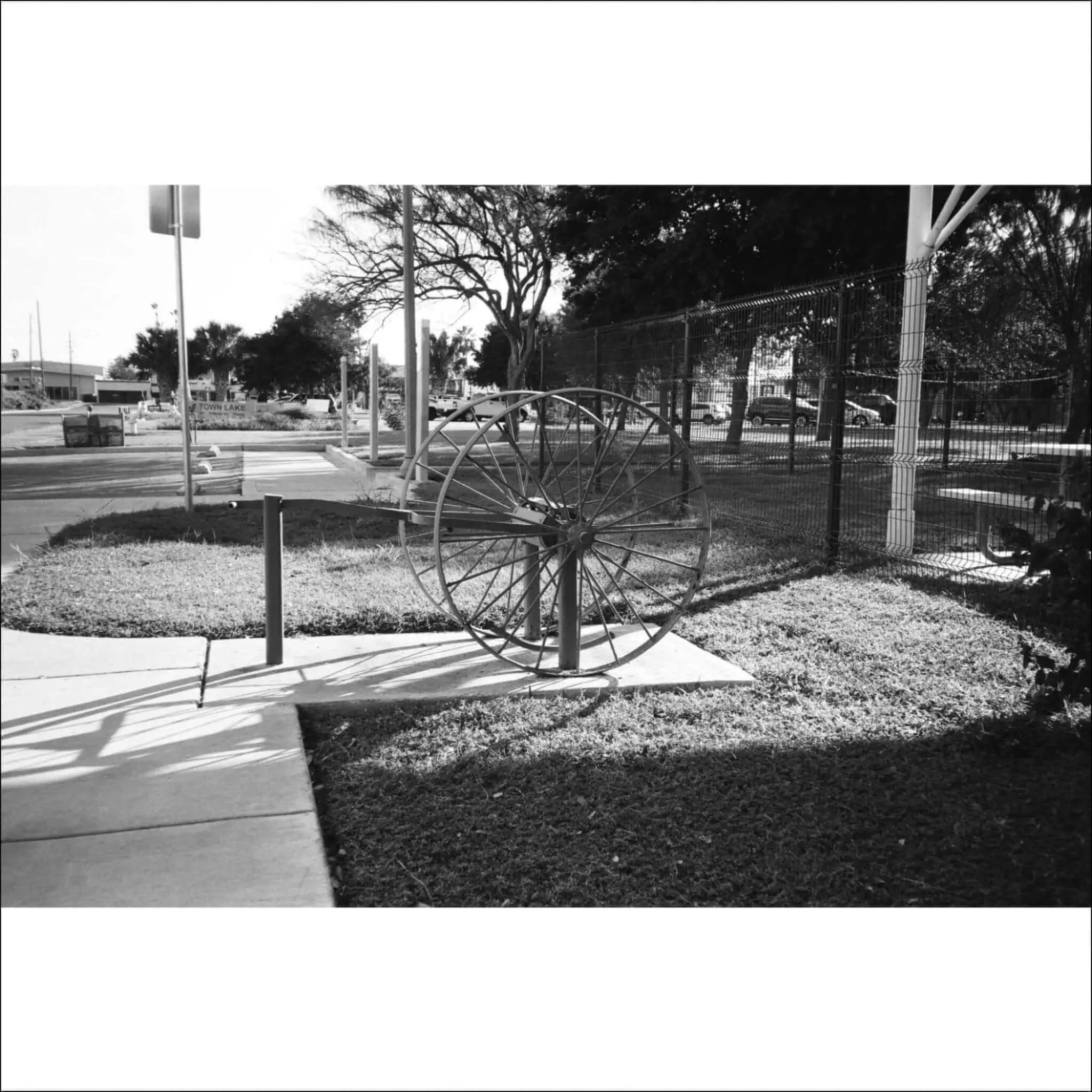Black and white photograph of a vintage cart wheel resting on grass near a sidewalk.
