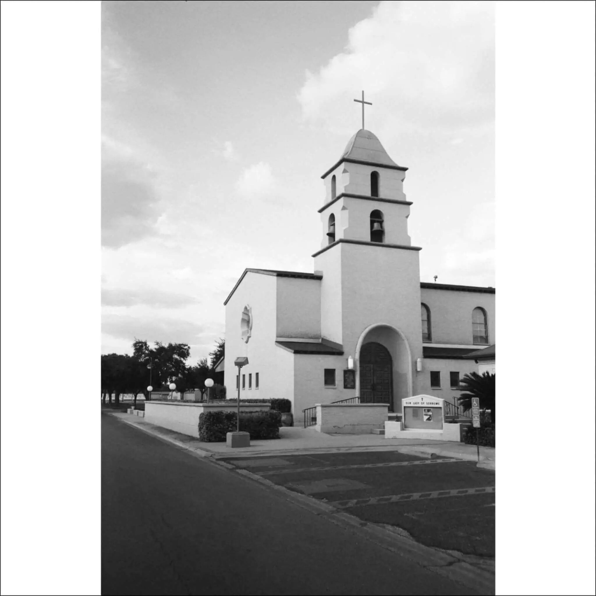Black and white photograph of a church building with a cross, sidewalk, and trees.