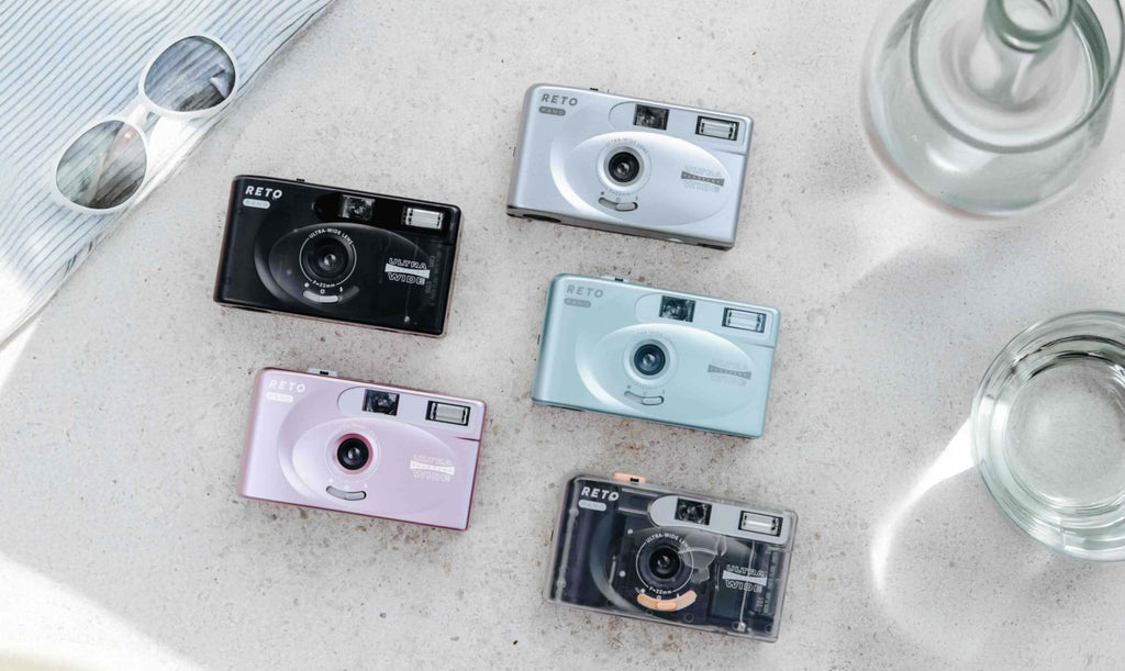 Five compact reto brand instant cameras in black, silver, light blue, pink and transparent with orange accents.