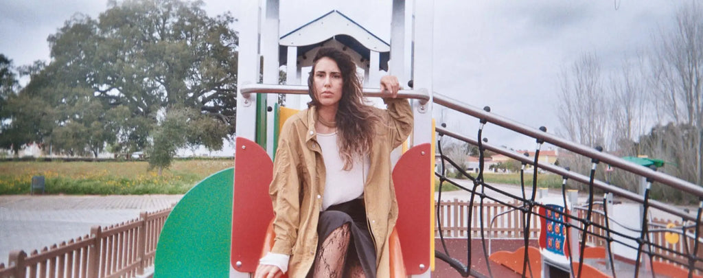 A woman with long, wavy brown hair stands on a colorful playground structure wearing an oversized tan jacket over a white top and patterned leggings.
