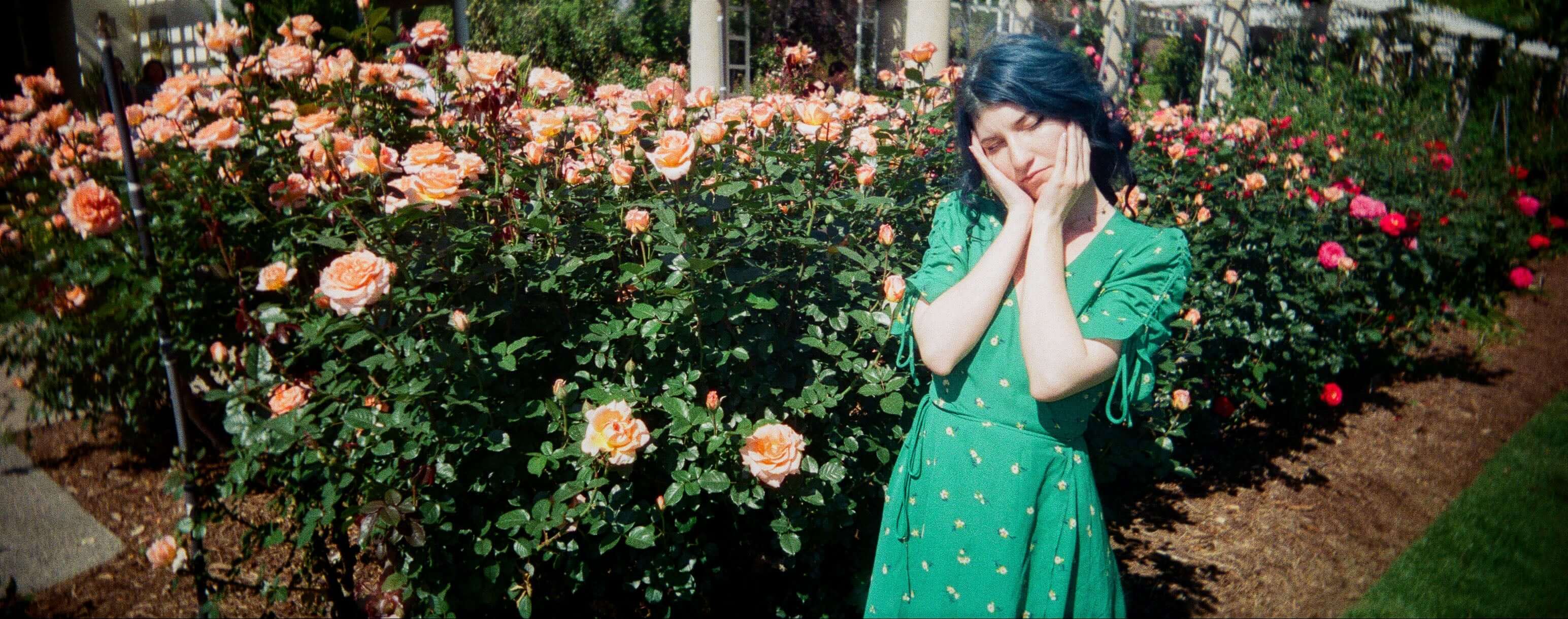 Young woman in a green dress posing amidst blooming roses in a sunny garden.