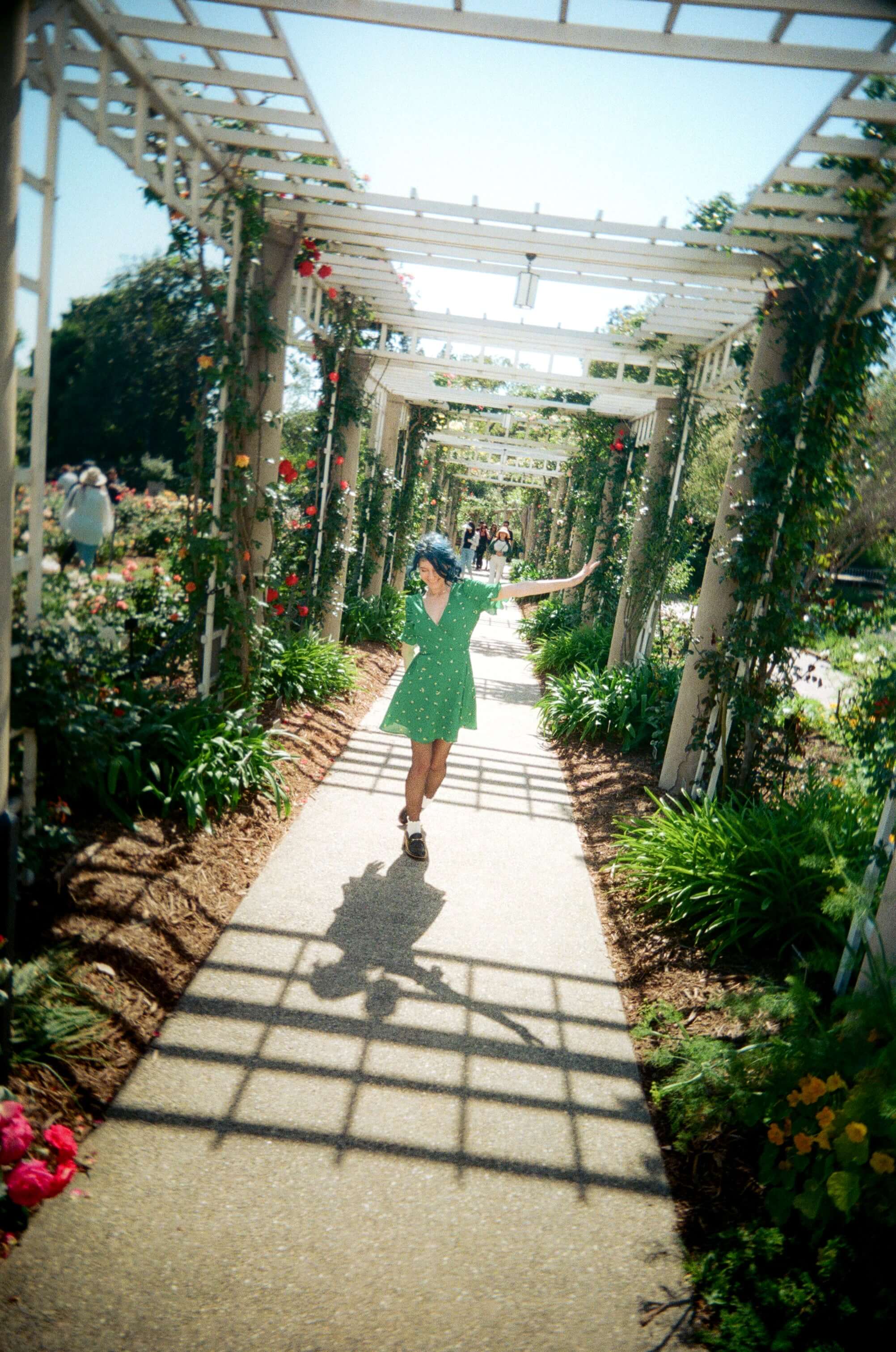 A joyful woman in a green dress dancing down a sunlit garden path adorned with flowers and greenery.