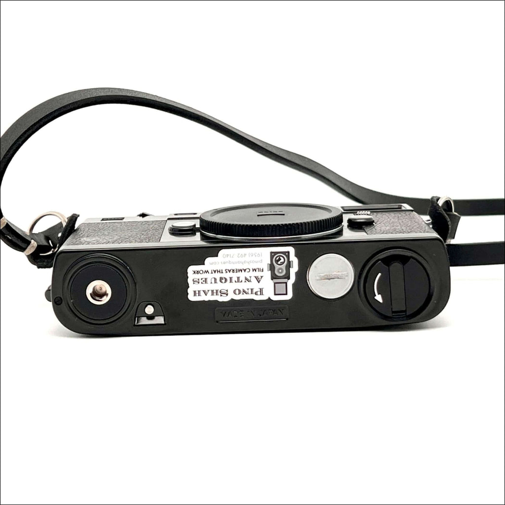 Black textured body with a silver shutter button and black leather strap, featuring a sticker that reads ’pino shah antiques film cameras that work.’