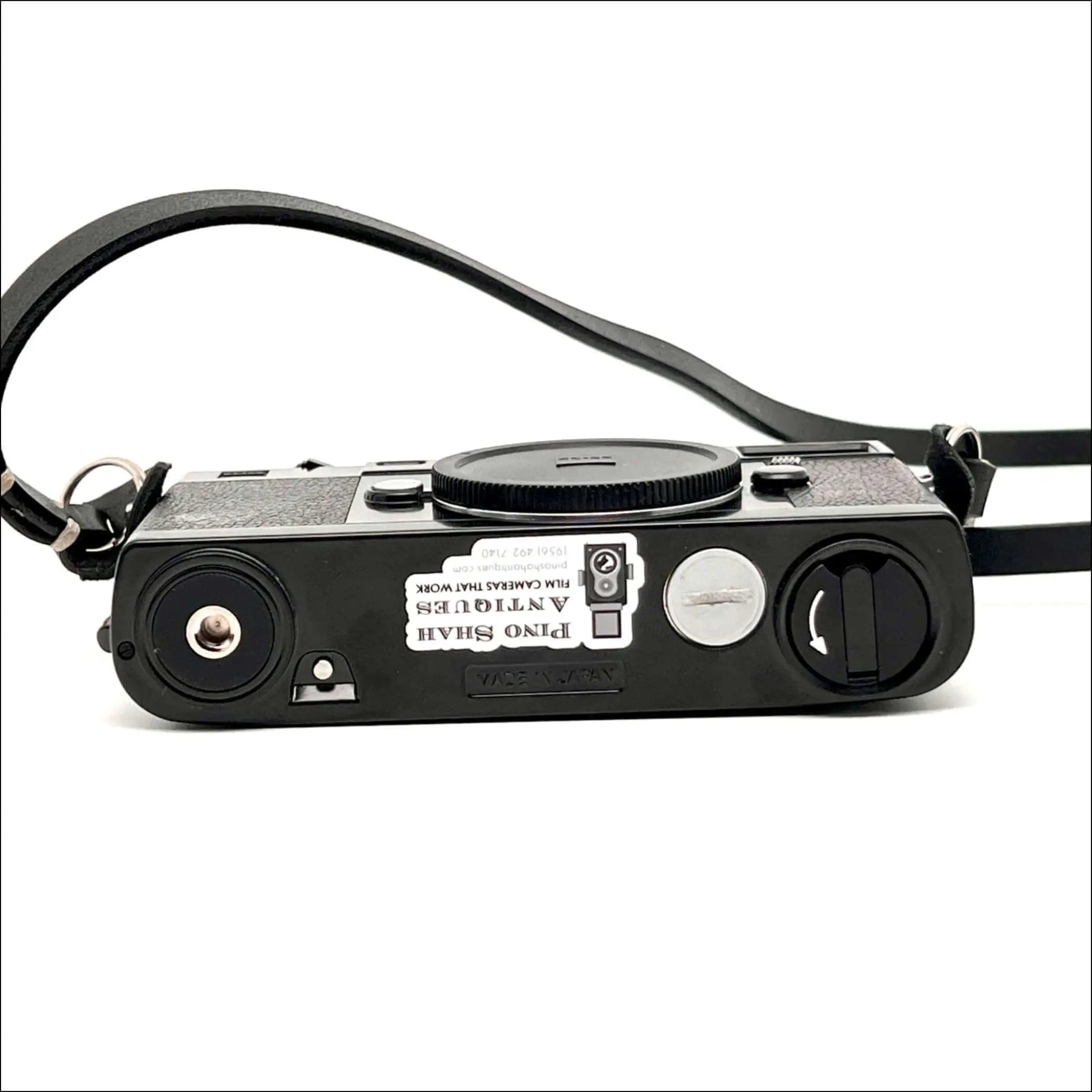 Black textured body with a silver shutter button and black leather strap, featuring a sticker that reads ’pino shah antiques film cameras that work.’