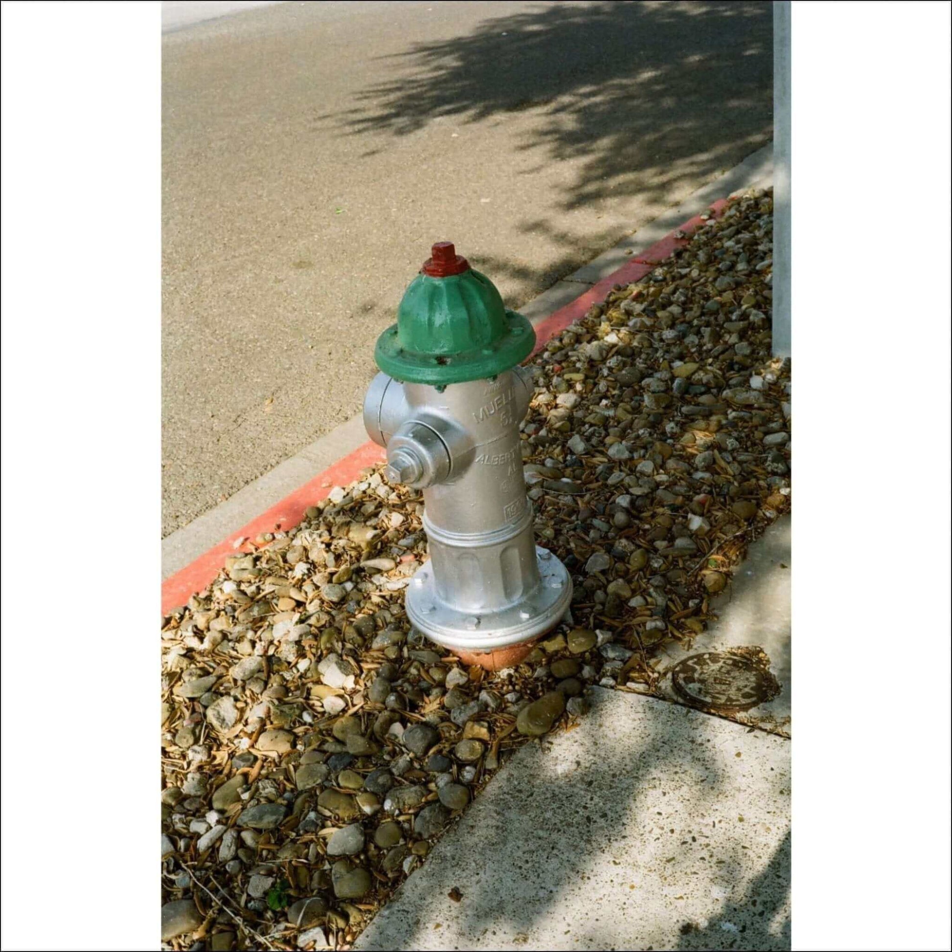 Silver and green fire hydrant on a gravel path under sunlight.