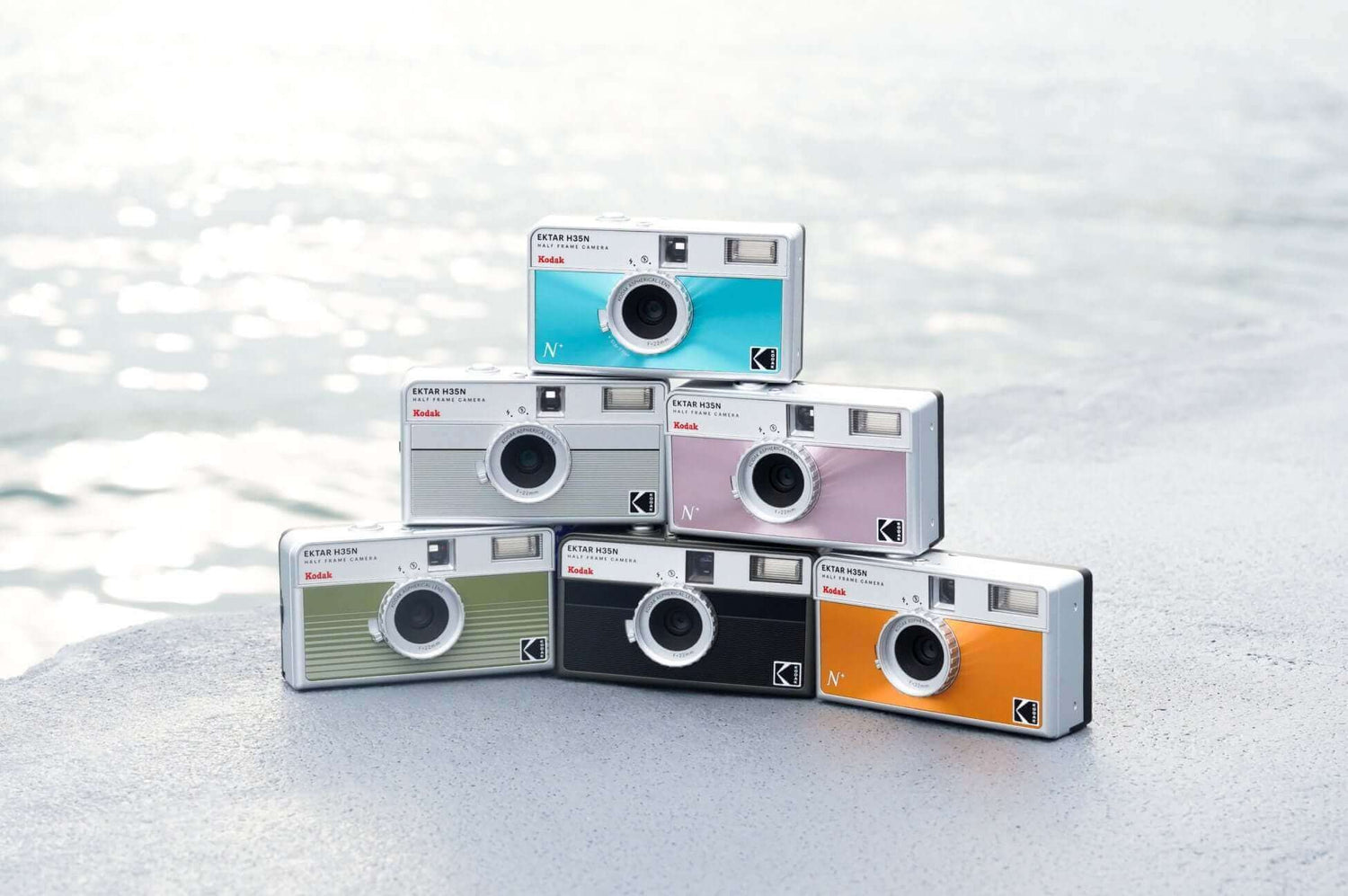 Colorful Kodak Ektar H35N Half-Frame cameras stacked by the water, showcasing stylish designs and features for photographers.