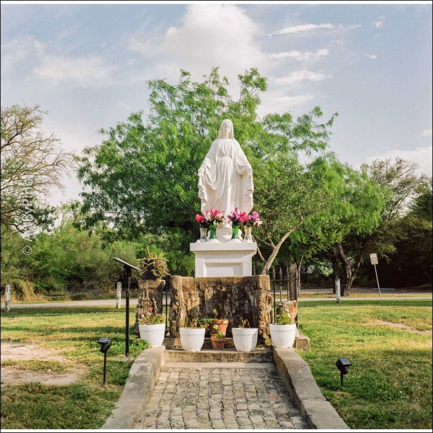Statue of a religious figure surrounded by flowers and trees at a serene outdoor location.