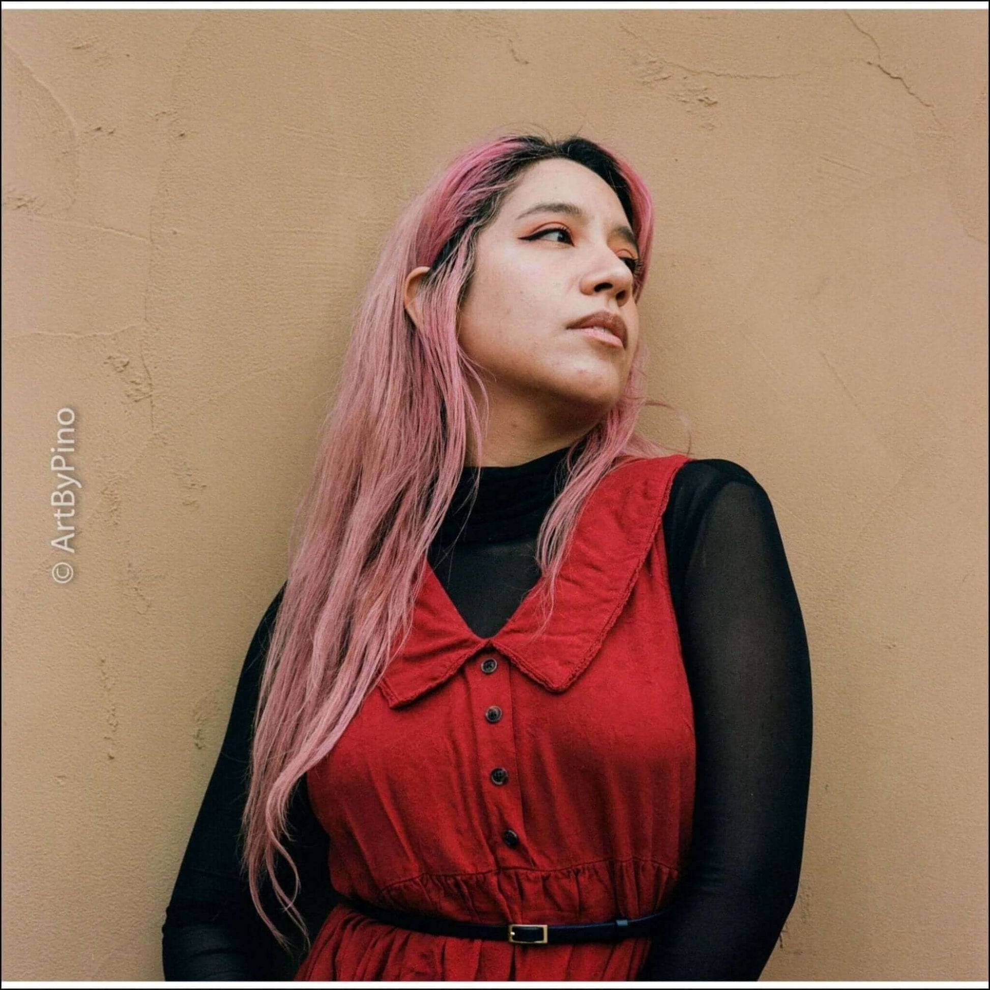 Portrait of a woman with pink hair wearing a red dress against a neutral background, conveying a stylish and artistic vibe.